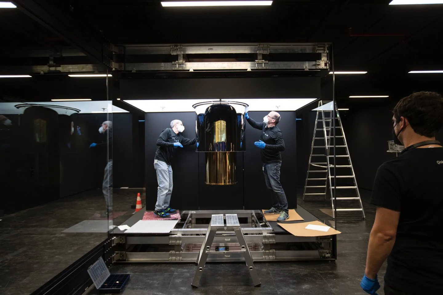 Ingenieurs werken in een donkere zaal aan een kwantumcomputer: een hoge, goudkleurige cilinder achter glas.