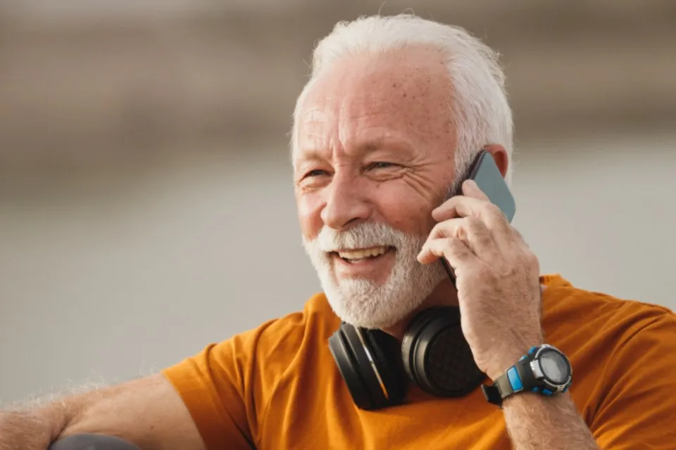 Een lachende oudere man met wit haar en baard houdt een telefoon aan zijn oor.