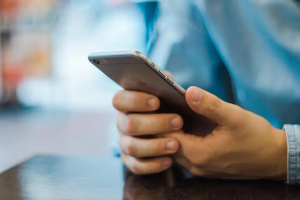 Twee handen houden een telefoon vast boven een tafel; de achterkant van het toestel is in beeld.